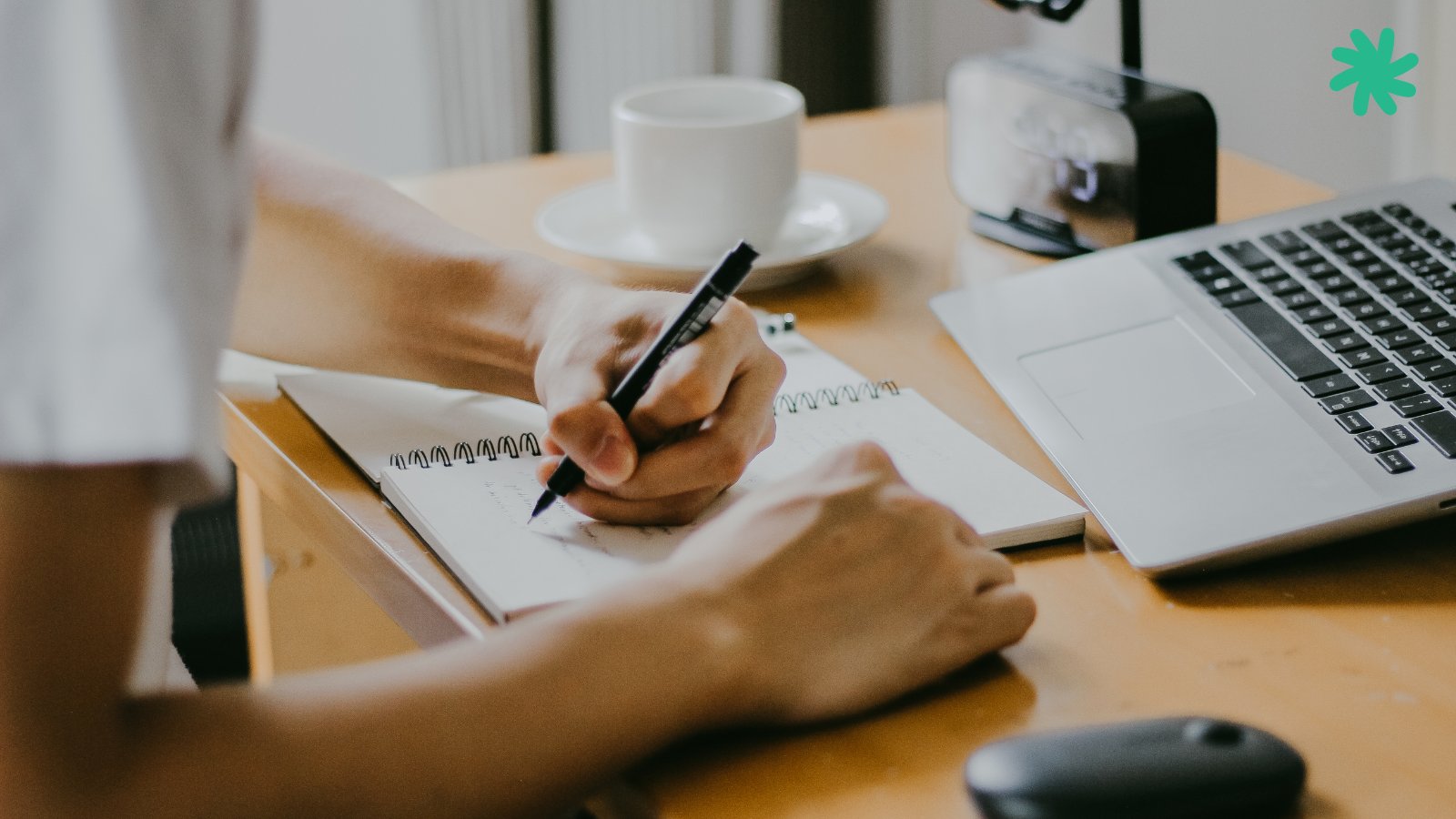Mãos escrevendo num caderno em mesa com notebook e xícara de café