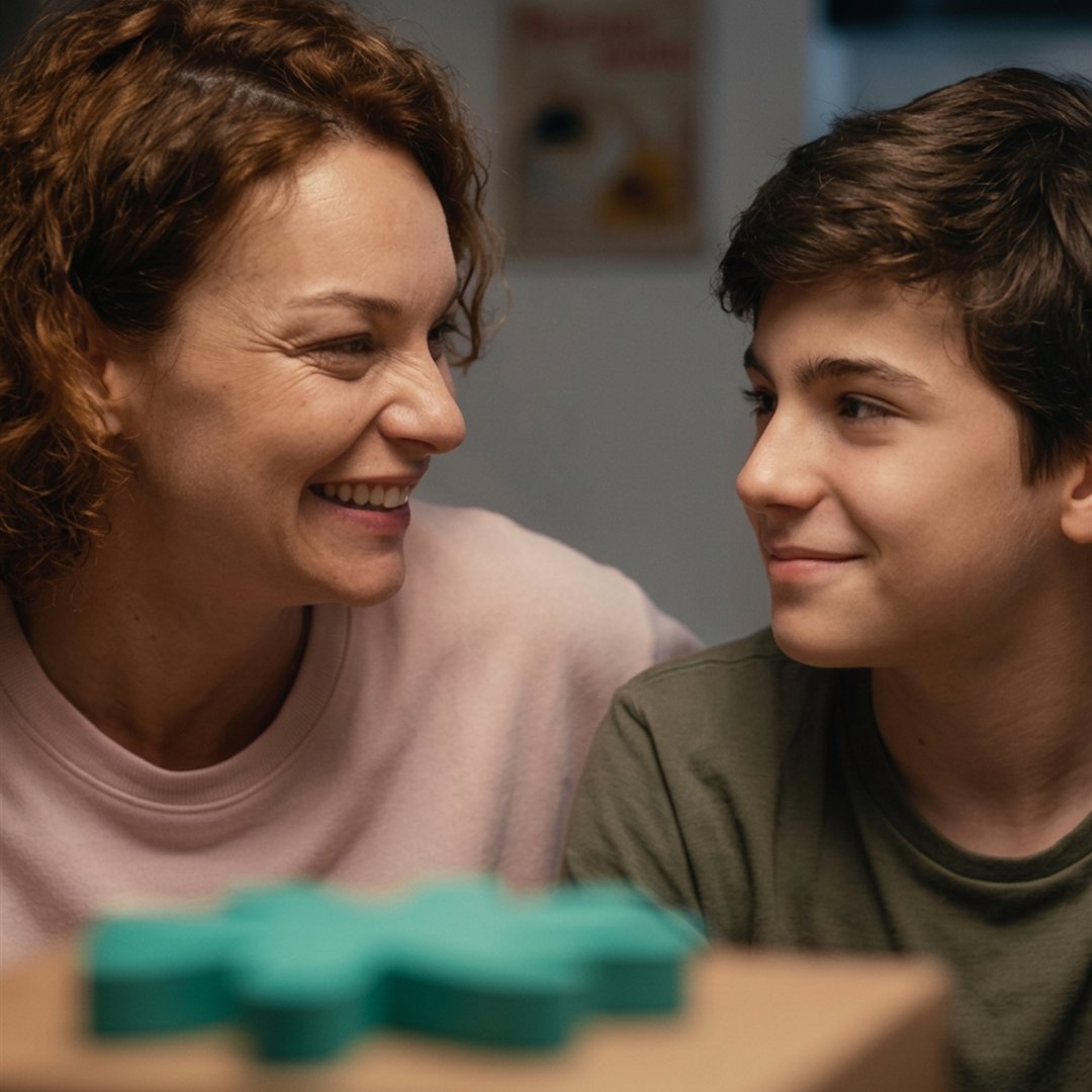 Mãe e filho adolescente lado a lado, sorrindo, em um momento de estudo em casa